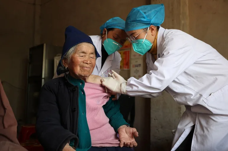 O femeie în vârstă primește o doză de vaccin COVID-19 de la un lucrător din domeniul sănătății.  Femeia pare relaxată.  Poartă un pulover roz, o haină neagră și o șapcă neagră.