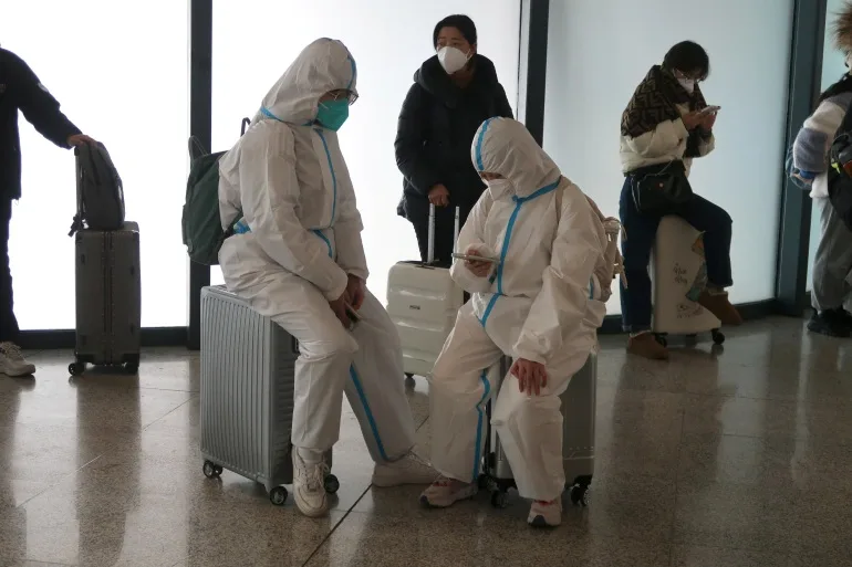 Călătorii îmbrăcați în costume albe pentru materii nocive stau pe bagaje în timp ce așteaptă să se îmbarce într-un tren.  Unul se uită la telefonul lor.