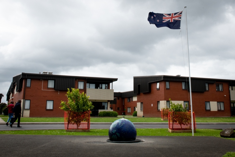 Centrul de Relocare a Refugiaților Mangere din Auckland, Noua Zeelandă.  Aceasta este o clădire din cărămidă roșie cu două etaje, cu un acoperiș maro și balcoane.  Un steag Noua Zeelandă flutură, iar grădina este curată și ordonată.  Două persoane merg spre clădire cu pungi de cumpărături