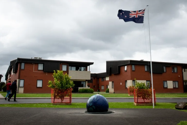 Centrul de Relocare a Refugiaților Mangere din Auckland, Noua Zeelandă.  Aceasta este o clădire din cărămidă roșie cu două etaje, cu un acoperiș maro și balcoane.  Un steag Noua Zeelandă flutură, iar grădina este curată și ordonată.  Două persoane merg spre clădire cu pungi de cumpărături