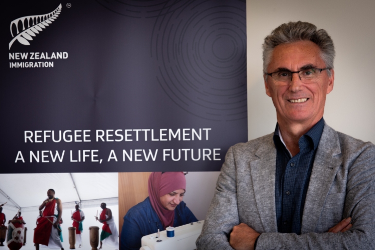 Un portret oficial al lui Qemajl Murati, managerul Programului de cote pentru refugiați pentru imigrarea în Noua Zeelandă, poartă o jachetă gri deschis și cămașă albastră cu gâtul deschis și zâmbește