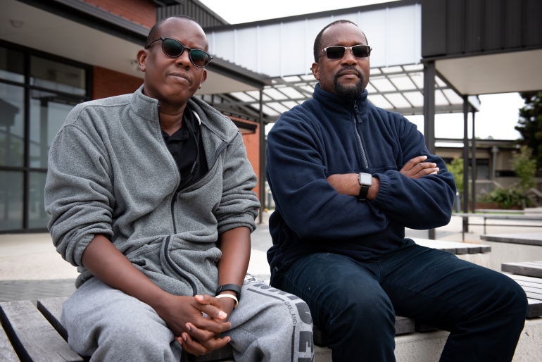 Mahmood din Somalia (stânga) și Jacques din Camerun (dreapta), stând pe o bancă în afara Centrului de Relocare a Refugiaților Mangere din Auckland.  Poartă lână și ochelari de culoare închisă și arată grijuliu.  Jacques are brațele încrucișate