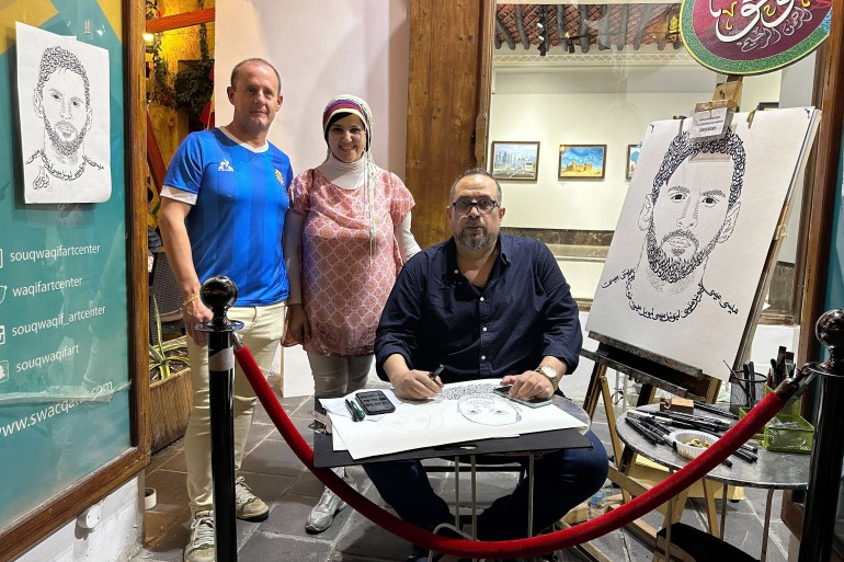 Khaled Almesawy stă cu o schiță sprijinită pe genunchi.  Există o schiță a lui Messi pe un șevalet în stânga lui și o alta atârnată pe perete în dreapta lui.  Două persoane pozează pentru o fotografie în spatele lui.