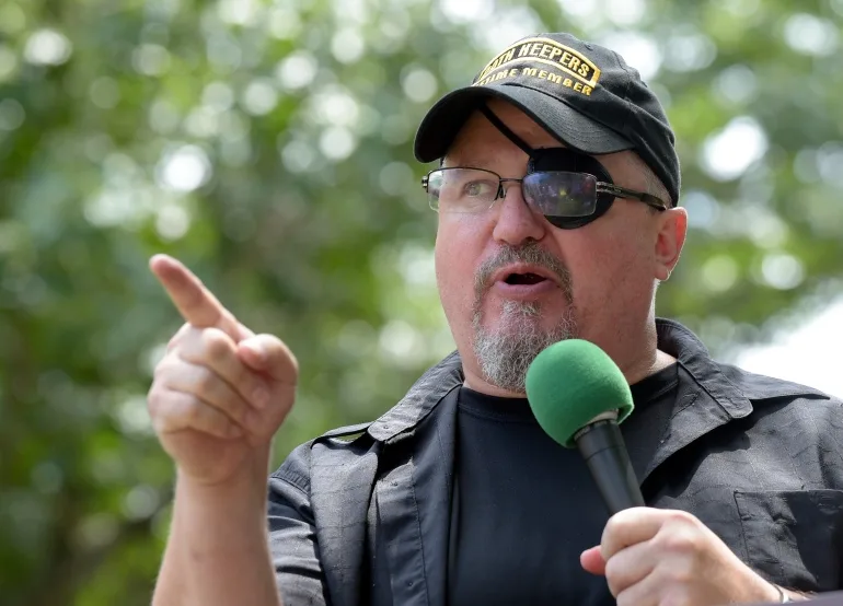 Stewart Rhodes, vorbind la microfon și arătând.  Poartă o șapcă neagră cu inscripții galbene Oath Keepers pe față.  Are și ochelari, care acoperă un plasture peste ochiul stâng.