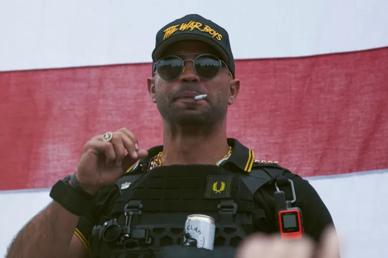 Enrique Tarrio, cu șapcă de baseball și vestă antiglonț.  Capacul negru are litere galbene care spun "BĂIEȚII RĂZBOIULUI.  Poartă ochelari de soare și are o țigară în gură.  Există o cutie de bere într-un suport pentru pahare atașat de jachetă.