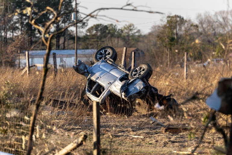 Un SUV răsturnat și distrus lângă un drum județean.  Se întinde printre iarbă uscată și crenguțe