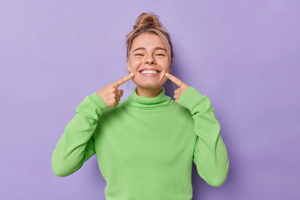 beautiful-happy-fair-haired-european-woman-points-index-fingers-mouth-forces-cheerful-smile-shows-perfect-white-teeth-wears-green-jumper-being-good-mood-isolated-purple-background